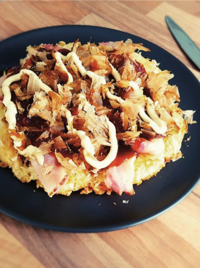 Assiette noire remplie d'okonomiyaki à la choucroute disposée sur une table en bois